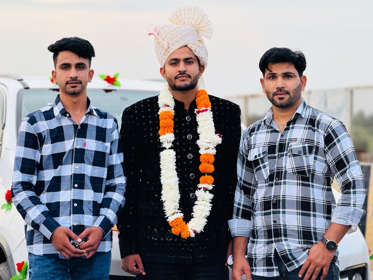 Three men pose outdoors at a cultural wedding, featuring traditional attire and floral garlands.