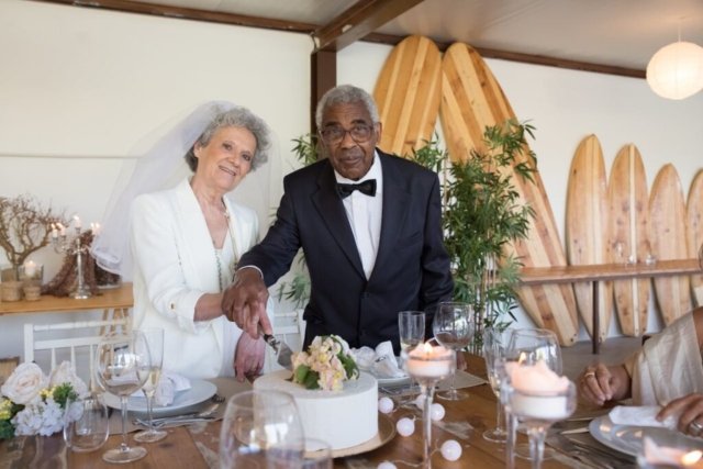 Senior couple elegantly cutting wedding cake indoors surrounded by floral decorations and candlelight.